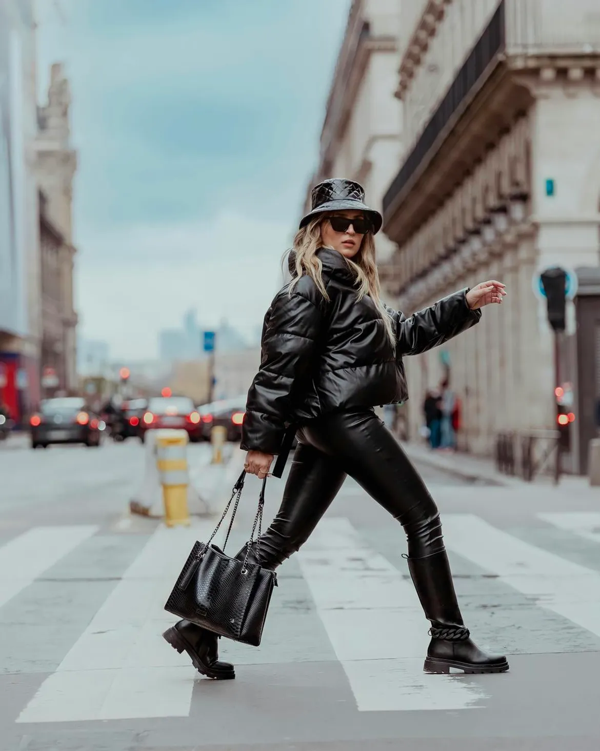 Woman in a black leather bucket hat, shiny leather puffer jacket, black leather pants, tote bag, and ankle boots walking on a Parisian street