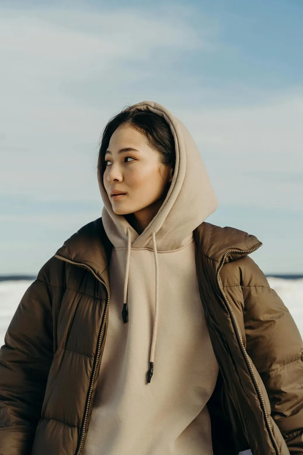 A woman dressed in a beige hoodie and brown puffer jacket outdoors in a winter setting.