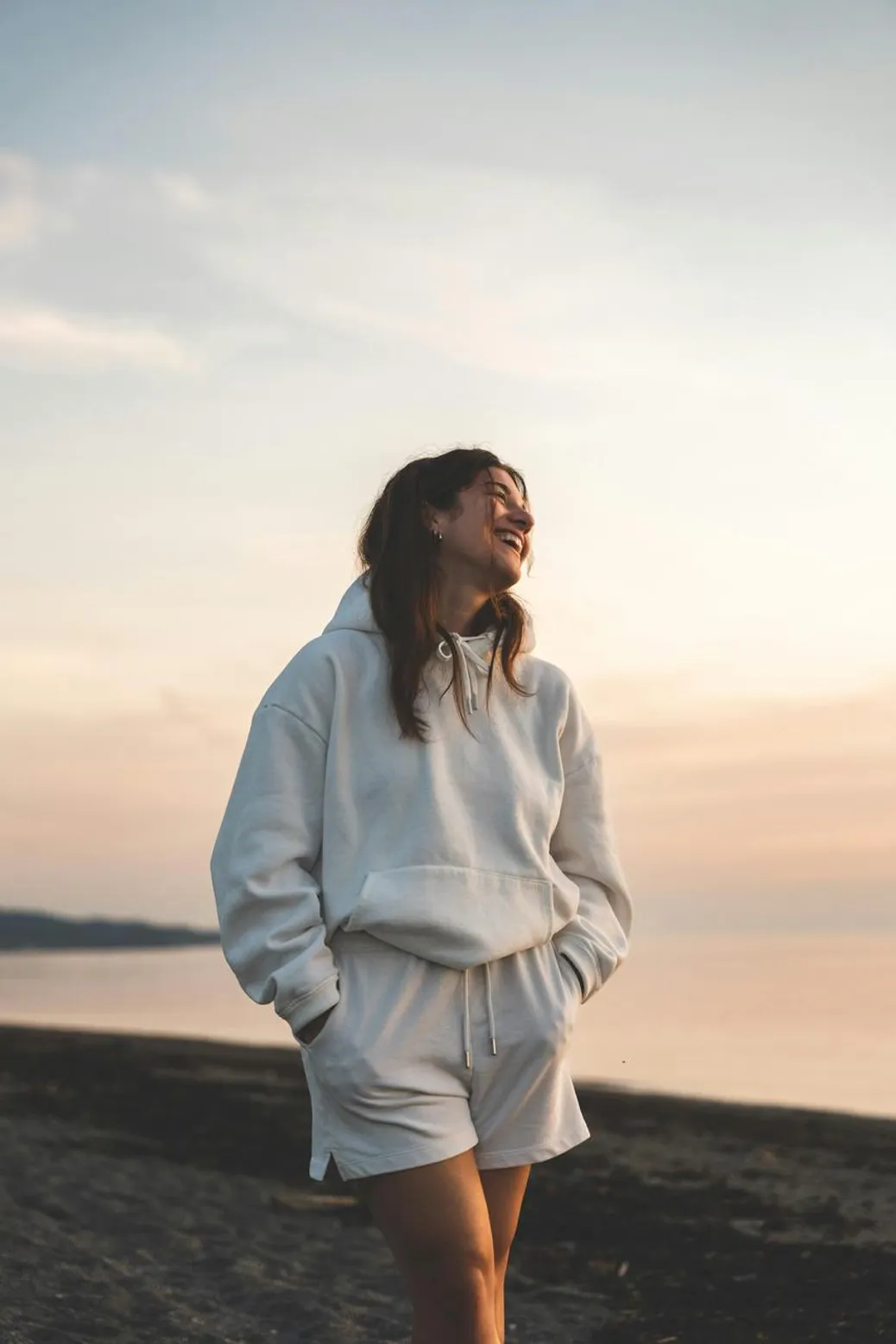 A woman in a white hoodie and shorts enjoying the beach at sunset.