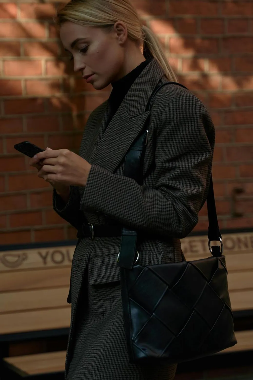 A stylish woman in a structured blazer checking her phone while carrying a black woven leather bag.