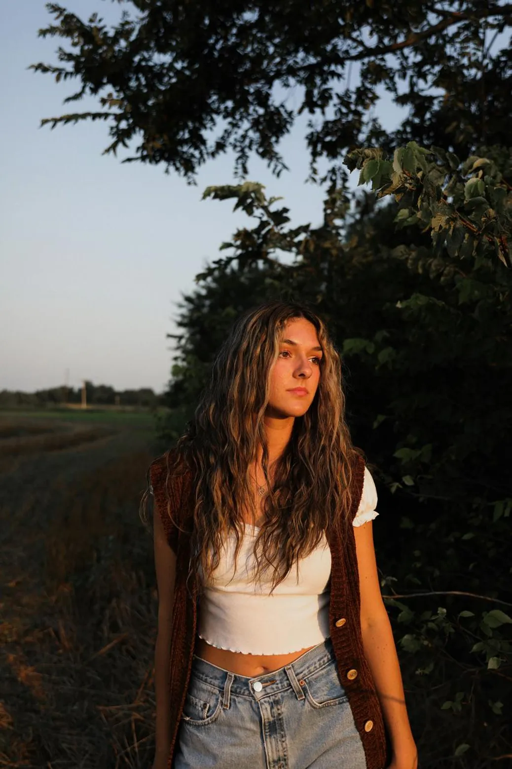 A woman with long wavy hair wearing a white crop top, knitted brown vest, and high-waisted denim jeans