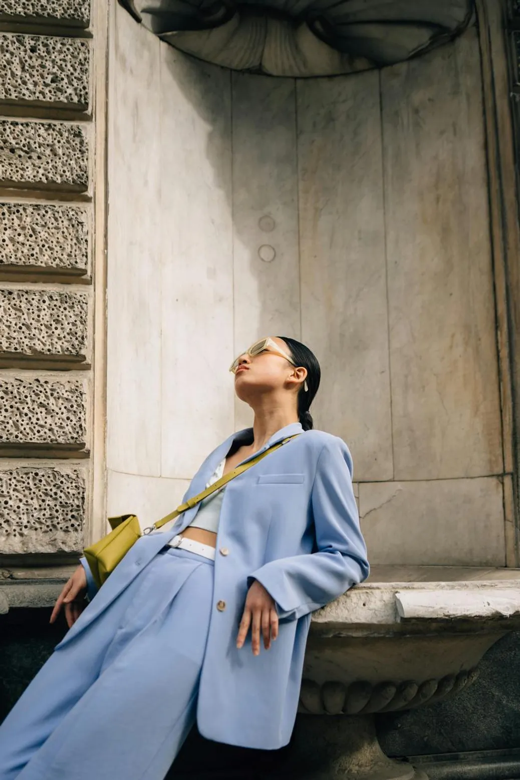 A stylish woman in a pastel blue suit leaning against a stone wall in an urban setting.