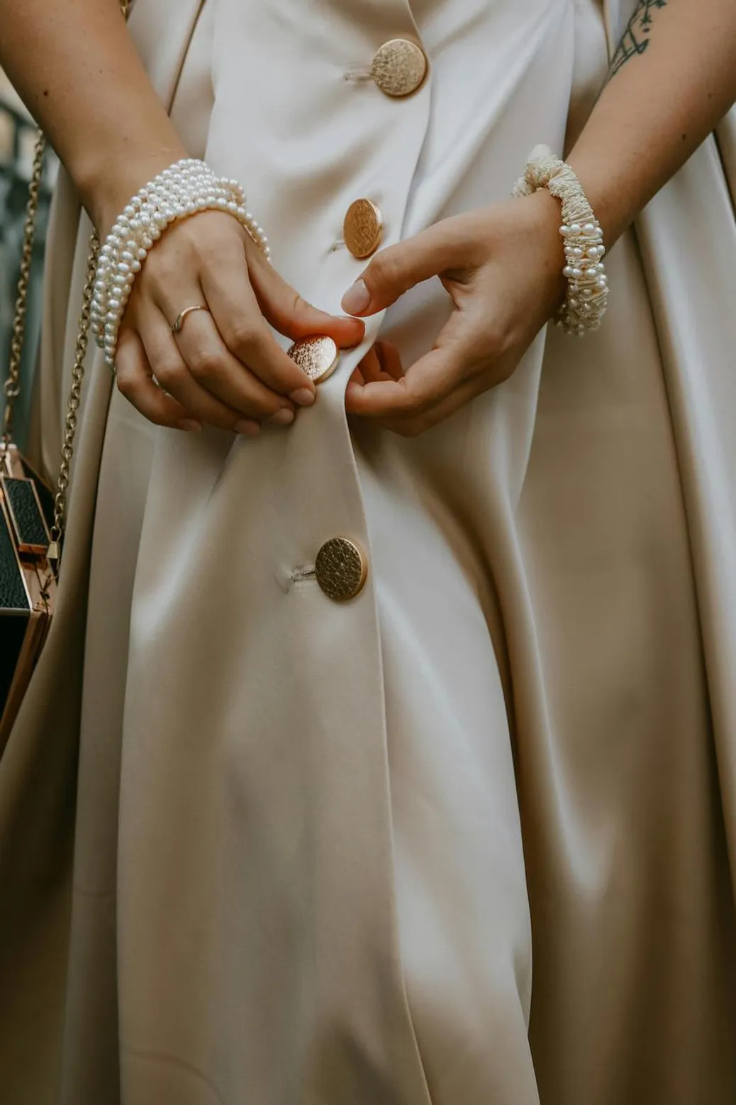 A close-up of a woman fastening elegant gold buttons on a satin dress, accessorized with pearl jewelry.